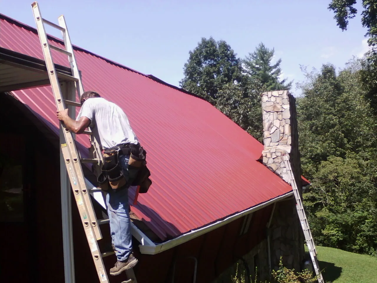 Roofer on a ladder beside a red metal roof and stone chimney — Soffit & Fascia Repair by Roofing Brothers in Ridgely
